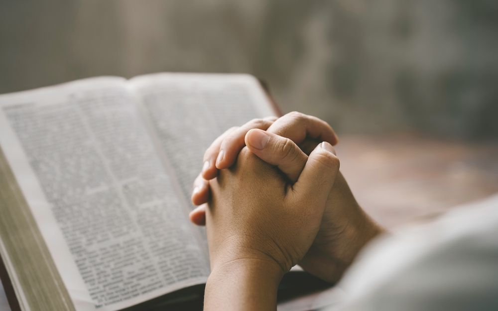 Hands clasped in prayer beside an open book on a wooden table