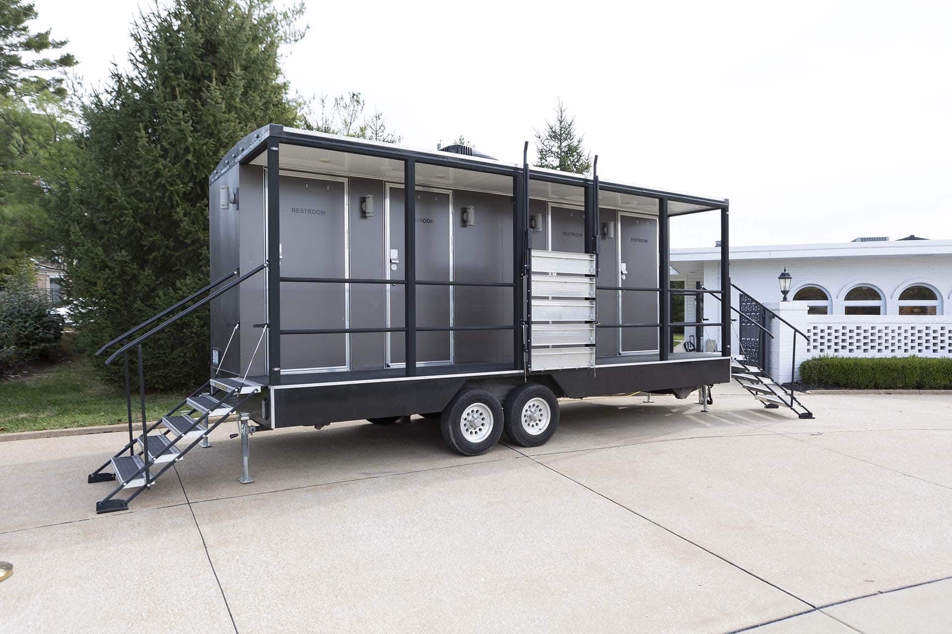 Gray portable restroom trailer on wheels with steps, parked on a concrete lot near trees and a building.