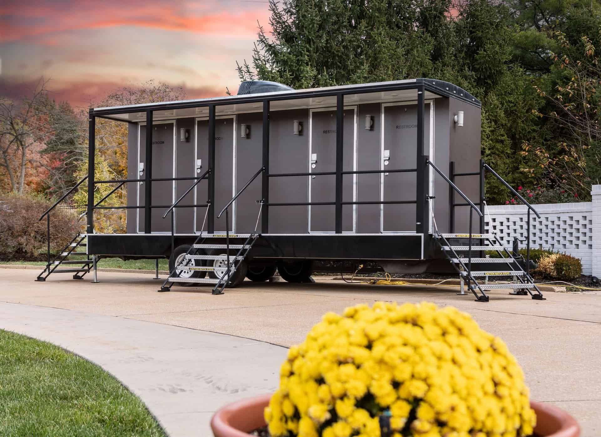 Gray portable building on a driveway, with stairs and railings, beside a yellow potted flower in the foreground.