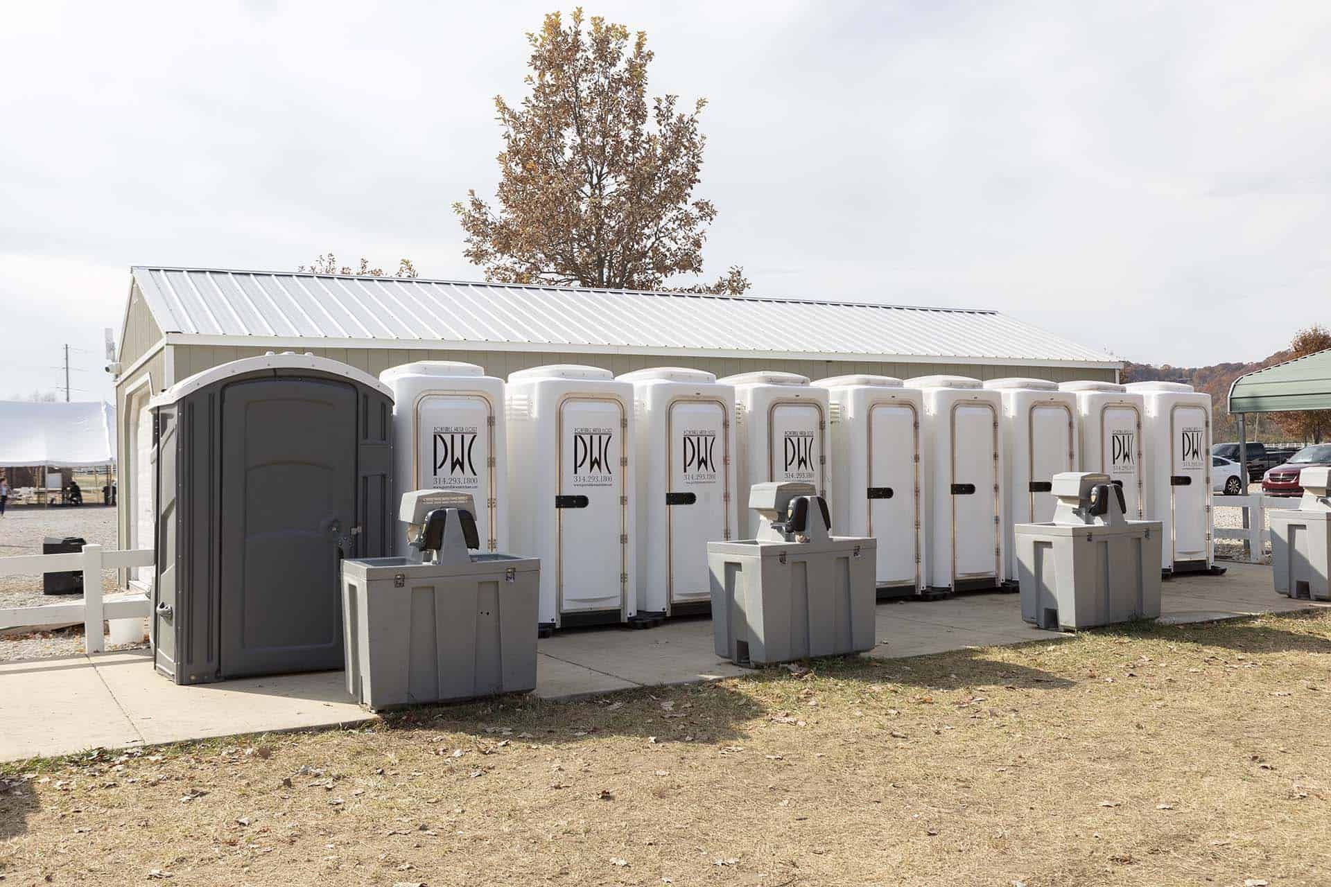 Portable restroom trailers lined up outdoors beside a white tent on a gravel lot.