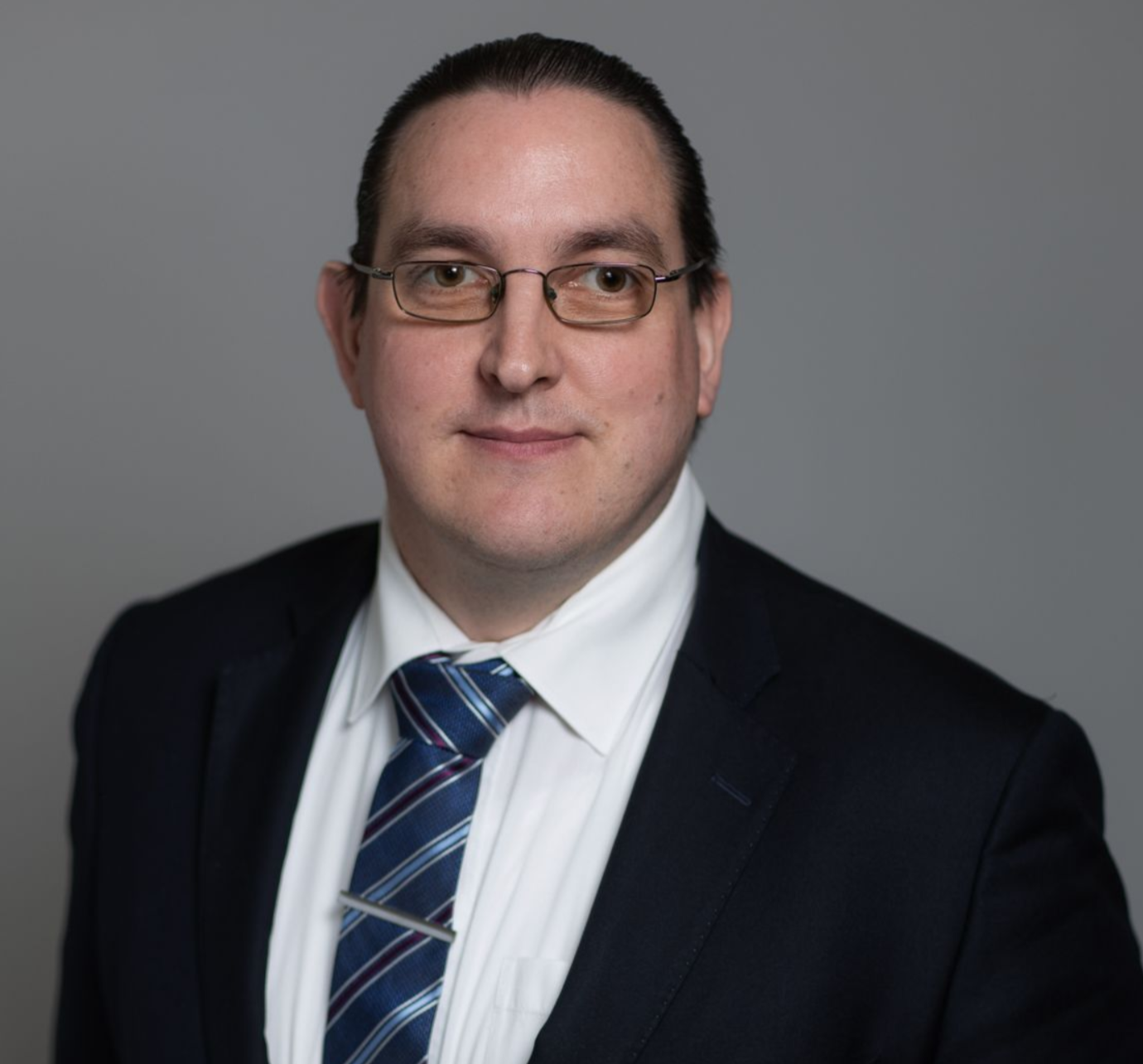 Studio headshot of a person in a dark suit, blue striped tie, and glasses against a gray background