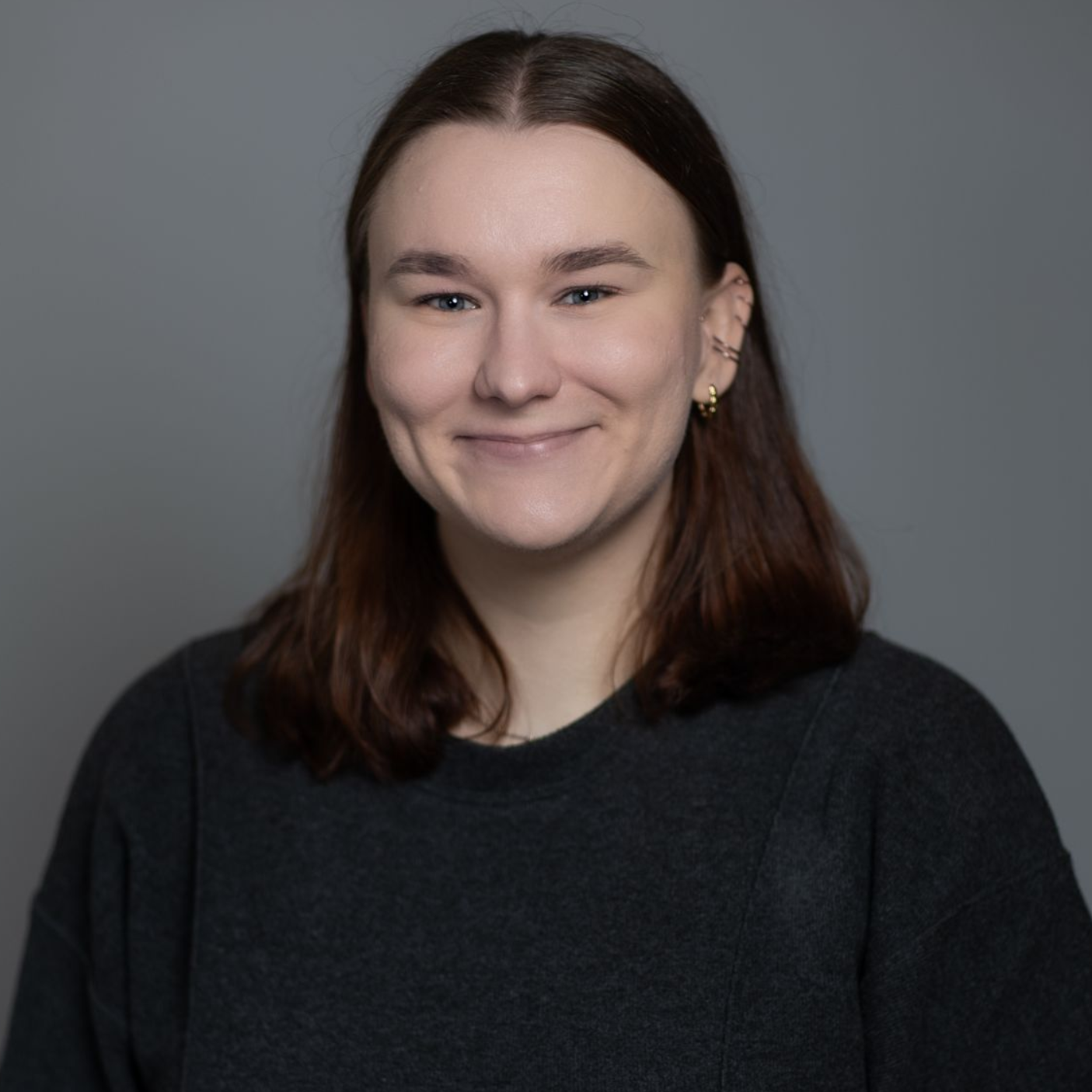 Portrait of a smiling person with shoulder-length brown hair against a gray background