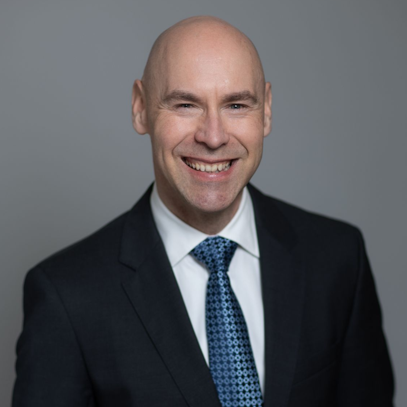 Smiling bald man in a dark suit and blue tie against a gray studio background
