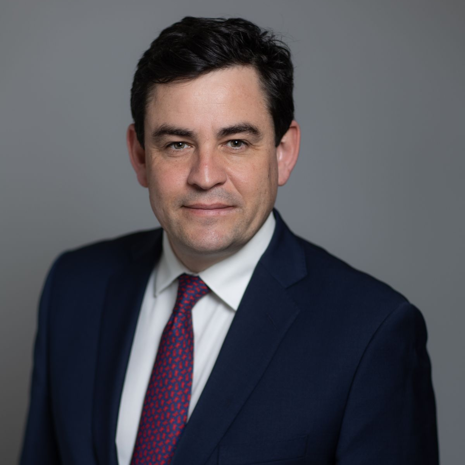 Studio portrait of a man in a navy suit, white shirt, and purple tie, facing forward with a neutral expression