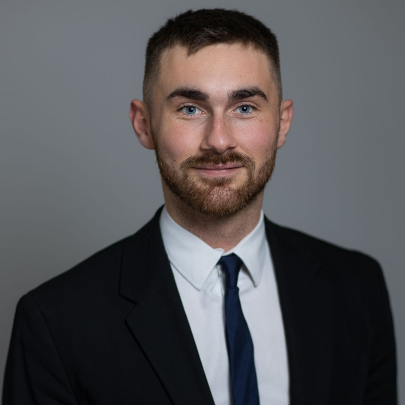 Studio headshot of a man in a black suit, white shirt, and blue tie against a gray background