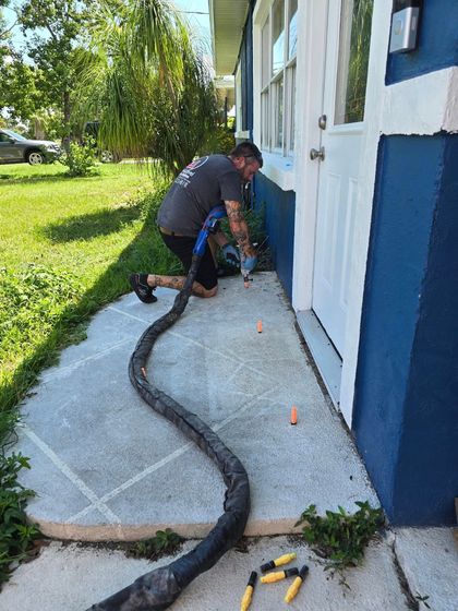 Worker installing a black hose along a white concrete walkway beside a blue-and-white building