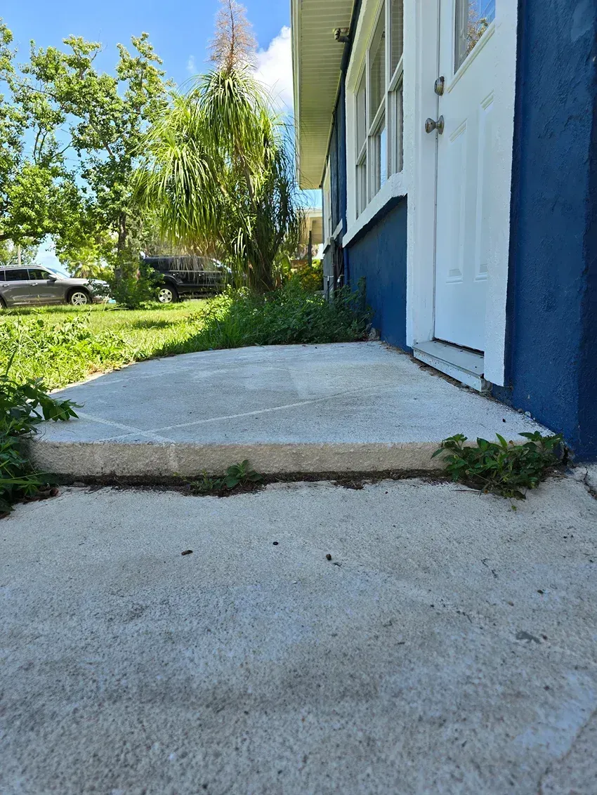 Concrete ramp and walkway beside a blue house, leading up to a front door with steps and grass nearby