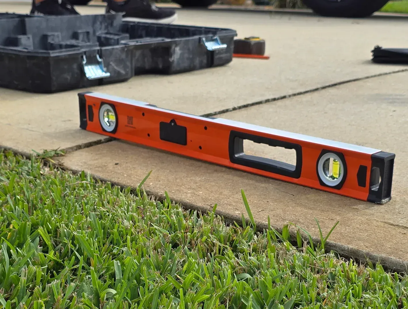Orange level tool on a sidewalk beside grass, with black equipment in the background.