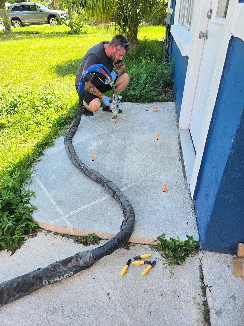 Worker crouching beside a blue house, repairing cracked concrete walkway with tools and orange markers.