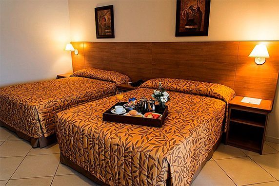 roupa de cama dourada estampada, uma bandeja de café da manhã em uma das camas e luminárias de parede com luz quente.