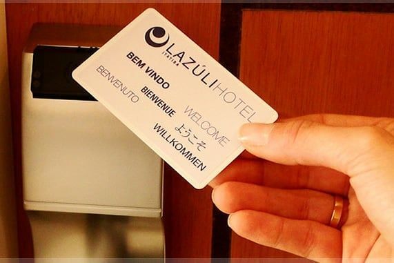 Uma mão segurando o cartão-chave de um quarto do Hotel Lazuli em frente a uma fechadura eletrônica da porta do hotel.