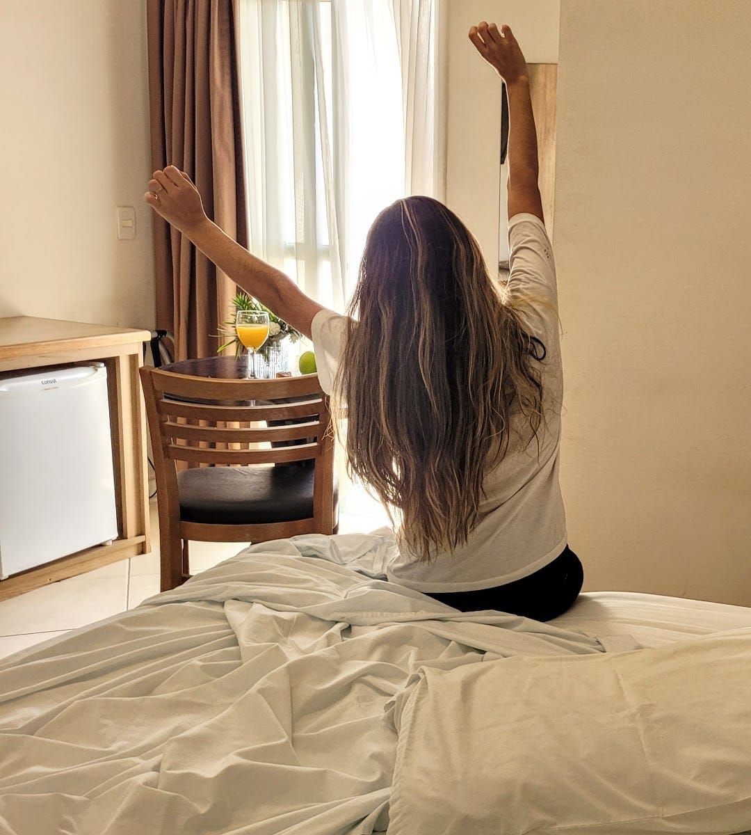 Uma mulher de cabelos compridos está sentada em uma cama, estendendo os braços em direção à janela em um quarto no Lazuli