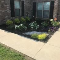A landscaped garden bed with flowering shrubs and a decorative stone sits against a brick house with black shutters.