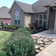 A brick house with a front garden featuring a stone walkway, green shrubs, and a small tree.