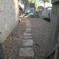 A stone stepping-stone path leads through a gravel side yard toward a backyard play structure.