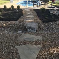 A stone pathway leads through a gravel yard toward a backyard swimming pool and patio furniture.