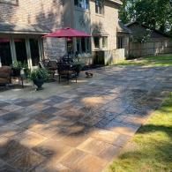 A stone patio with a red umbrella and outdoor furniture sits next to a brick house in a backyard.