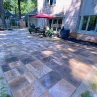 A large stone patio with a red umbrella, outdoor dining set, and grill outside a brick house.