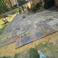 Two workers lay large, dark stone pavers in a residential backyard, using levels to ensure the patio is flat.