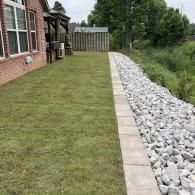 A brick house with a green lawn next to a stone-lined garden border and a white fence in a backyard.