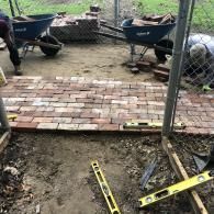 Two people build a red brick walkway outdoors, with two blue wheelbarrows and spirit levels nearby.
