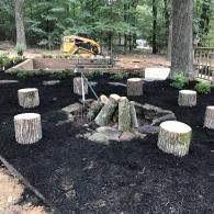 A fire pit surrounded by tree stump seats in a mulched backyard, with a construction vehicle in the background.