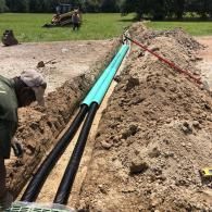A worker installs two black corrugated pipes and a green pipe into an open trench at a construction site.