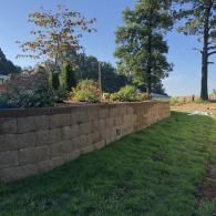 A beige, segmented concrete retaining wall stands next to a grassy lawn under a clear blue sky, with trees in the background.