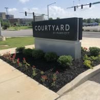 A black Courtyard by Marriott sign stands on a concrete base in a landscaped garden area by a street intersection.