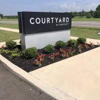 A black and white Courtyard by Marriott hotel sign stands in a landscaped garden bed with shrubs and flowers.