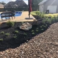 A backyard swimming pool area with a landscaped garden featuring dark mulch, small green plants, a rock, and river stones.
