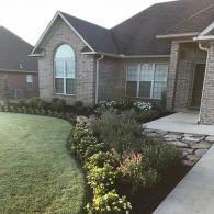 A brick house exterior with a curved front lawn, mulch beds, small shrubs, and a stone walkway leading to the entrance.