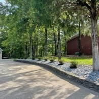 A paved driveway with a stone retaining wall and landscaped bed of shrubs running alongside a row of trees and a house.