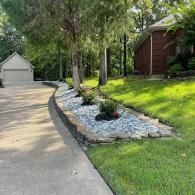 A concrete driveway leads to a garage next to a landscaped rock bed lined with stones, trees, and a grassy yard.