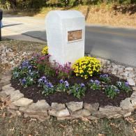 A white stone memorial marker sits in a garden bed surrounded by colorful flowers and bordered by stacked fieldstone.