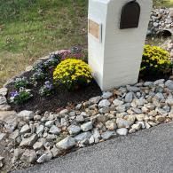 A white brick mailbox pedestal stands in a stone-bordered garden bed with yellow mums and small purple flowers.