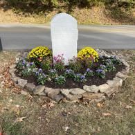 A blank, upright white headstone surrounded by yellow mums and purple pansies in a stone-bordered garden bed.