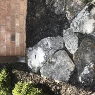 A close-up of light gray landscape rocks arranged next to a brick building wall, surrounded by dark mulch.