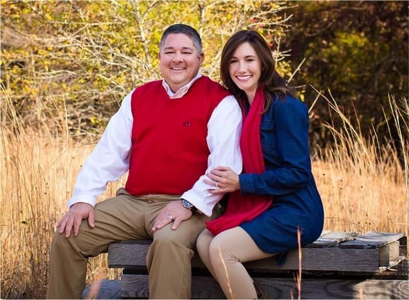 A couple sits together on a wooden bench outdoors against a background of dry grass and trees in autumn.
