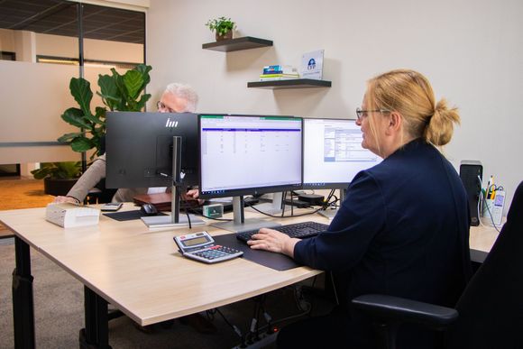 Twee kantoorpersoneel zit achter een bureau met beeldschermen en bekijkt spreadsheets.