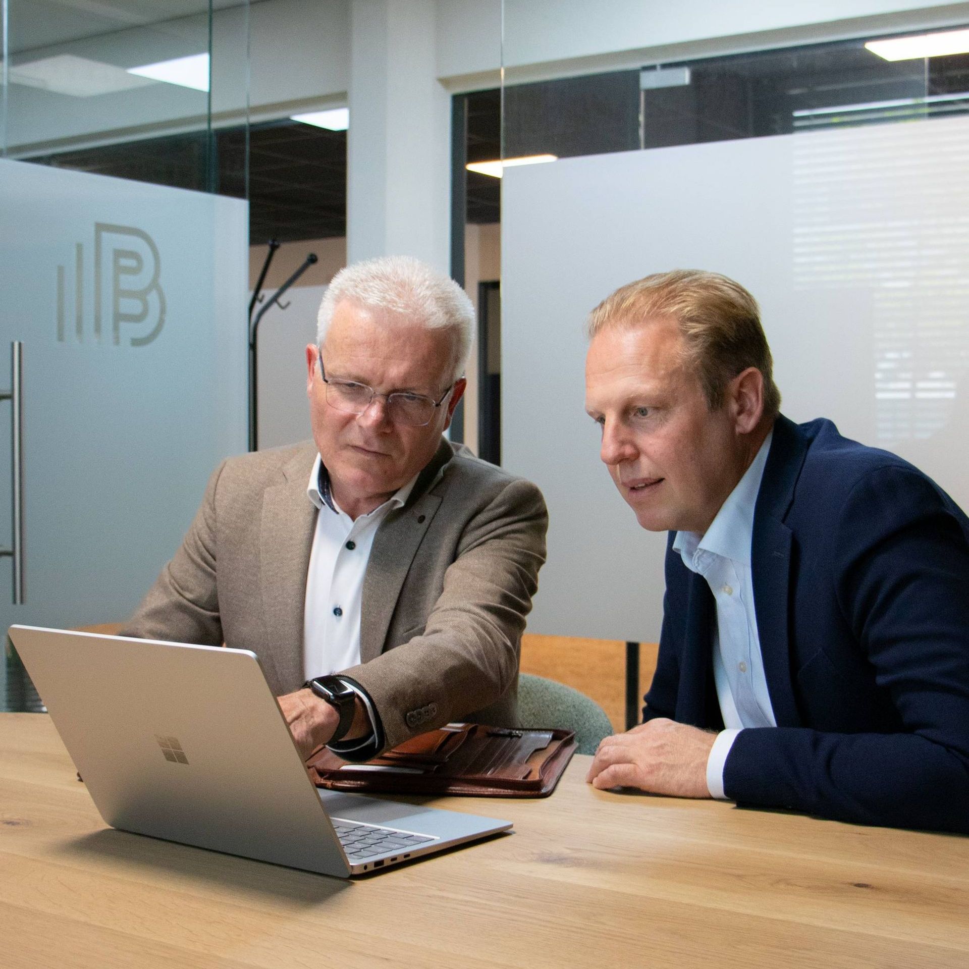 Twee mannen op kantoor kijken samen naar een laptop. De een draagt een bril en een bruin jasje. De ander een blauw jasje.