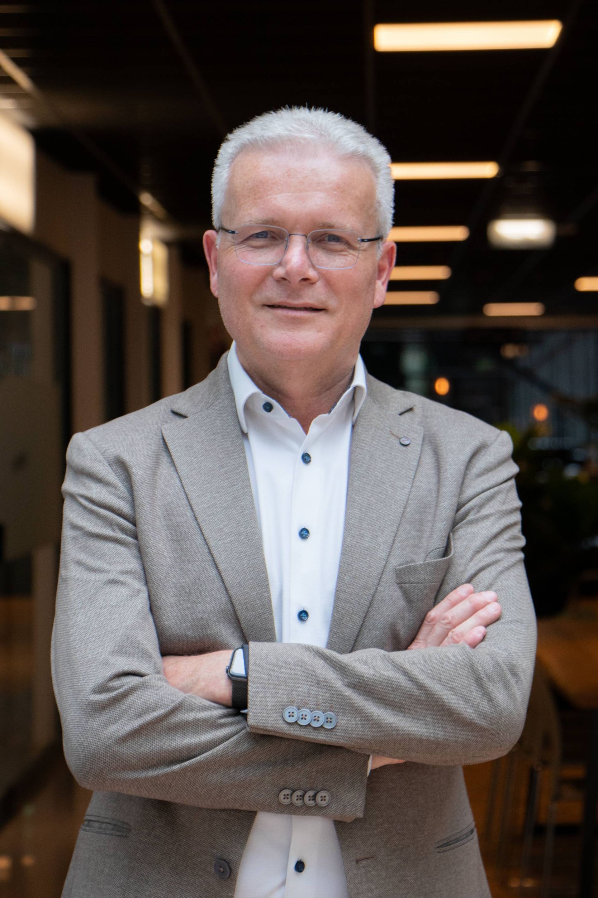 Man met wit haar en een bril, armen over elkaar, gekleed in een beige blazer en wit overhemd.