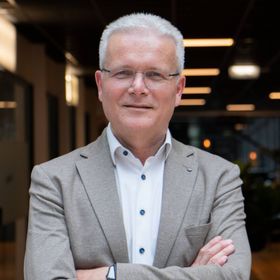 Man met grijs haar en een bril, armen over elkaar, gekleed in een lichtbruin jasje en wit overhemd, binnenshuis.