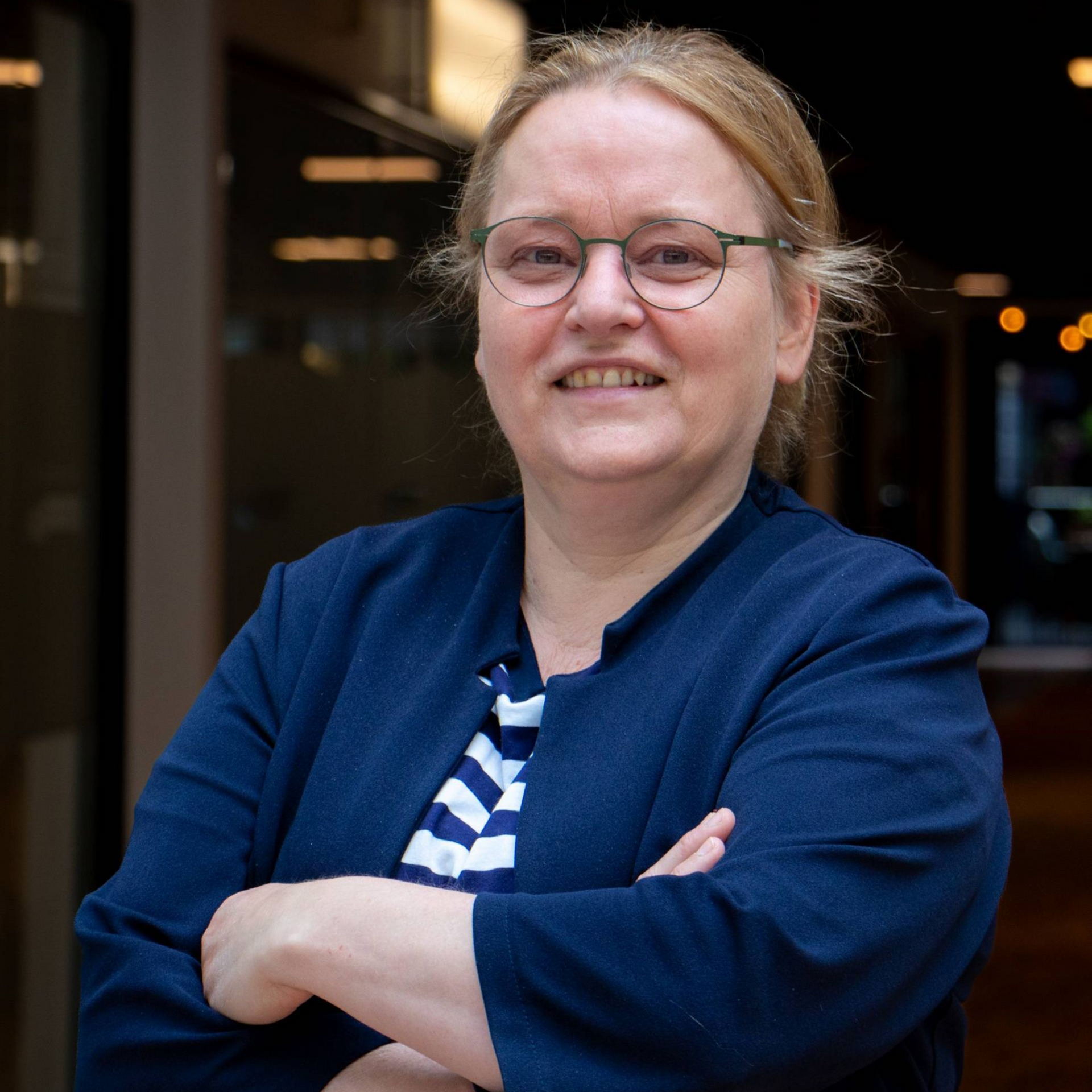 Vrouw met bril en gekruiste armen, glimlachend; draagt ​​een marineblauw jasje en een gestreept shirt.