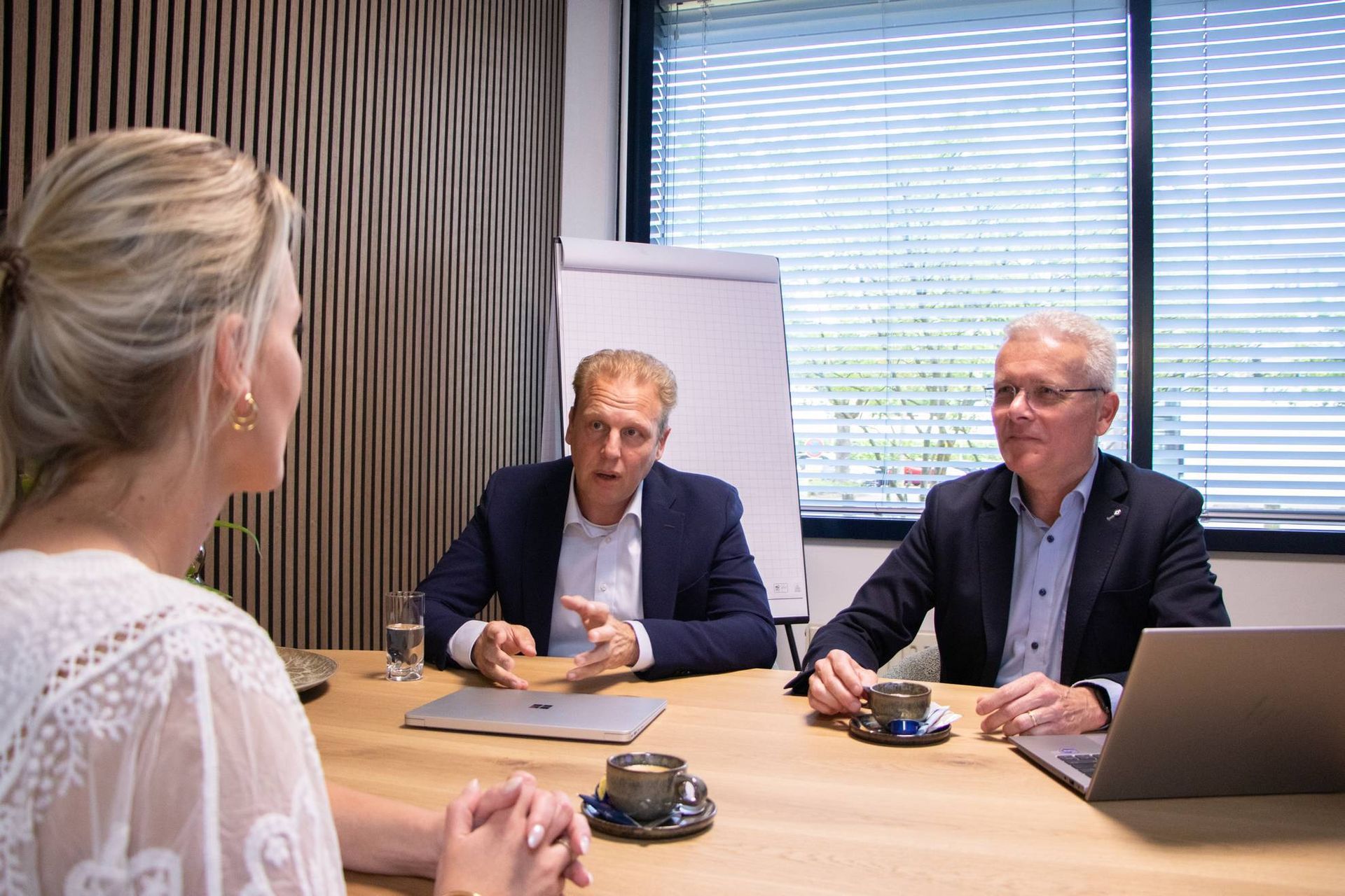 Drie mensen in vergadering: een vrouw tegenover twee mannen aan een tafel, kantooropstelling met jaloezieën.