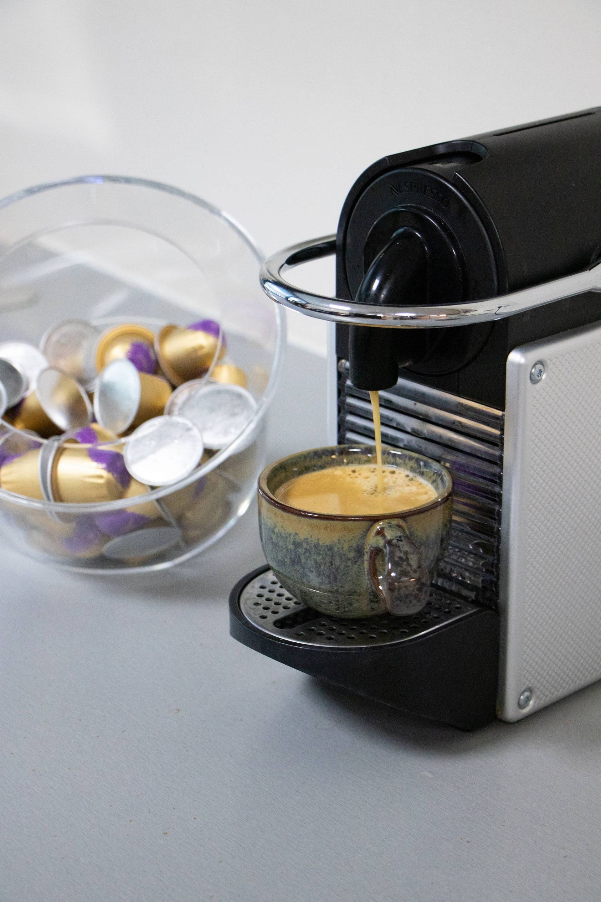 Espressomachine schenkt koffie in een klein mokje; capsules in een doorzichtig bakje aan de linkerkant.