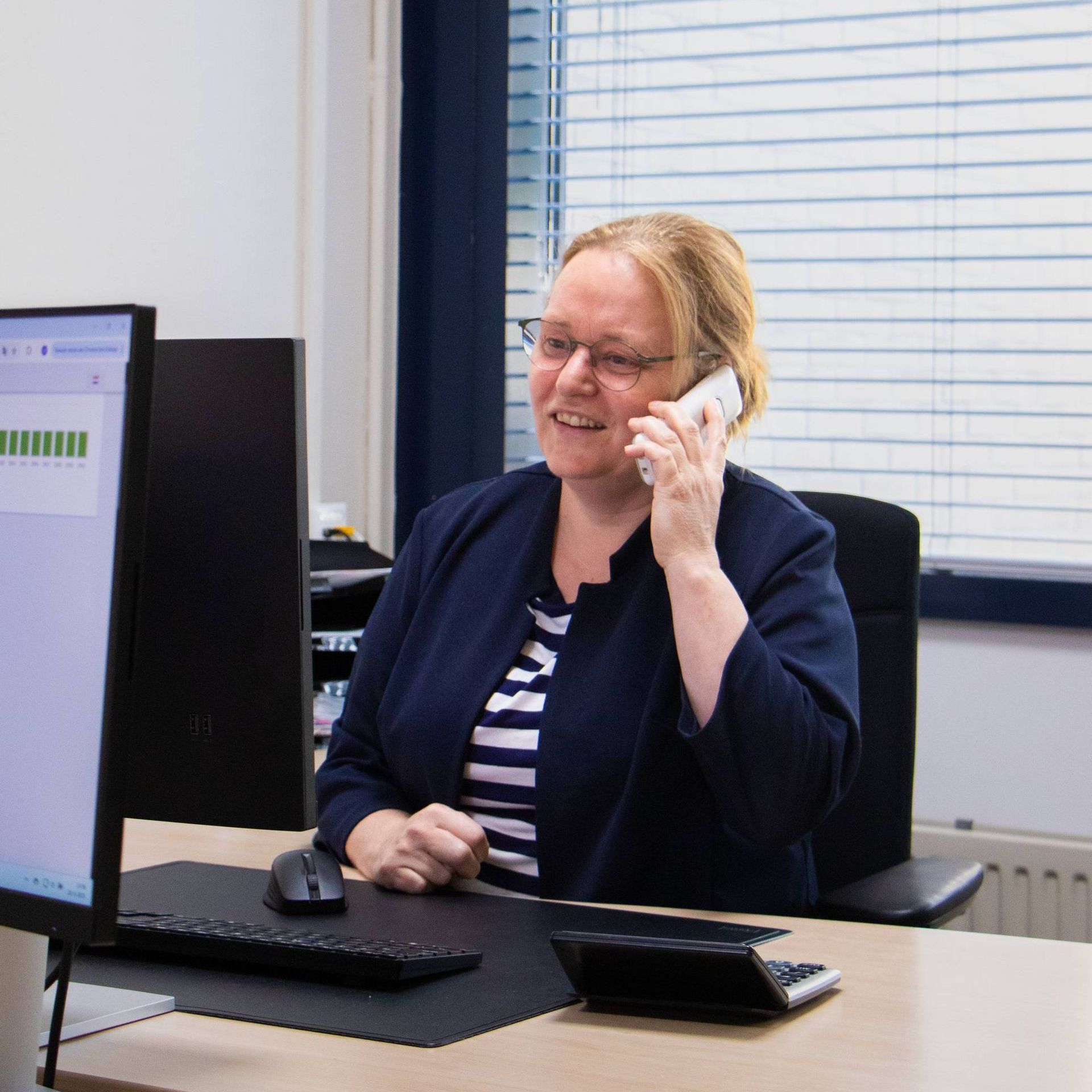 Vrouw met bril aan de telefoon, glimlachend aan bureau met computer en rekenmachine op kantoor.