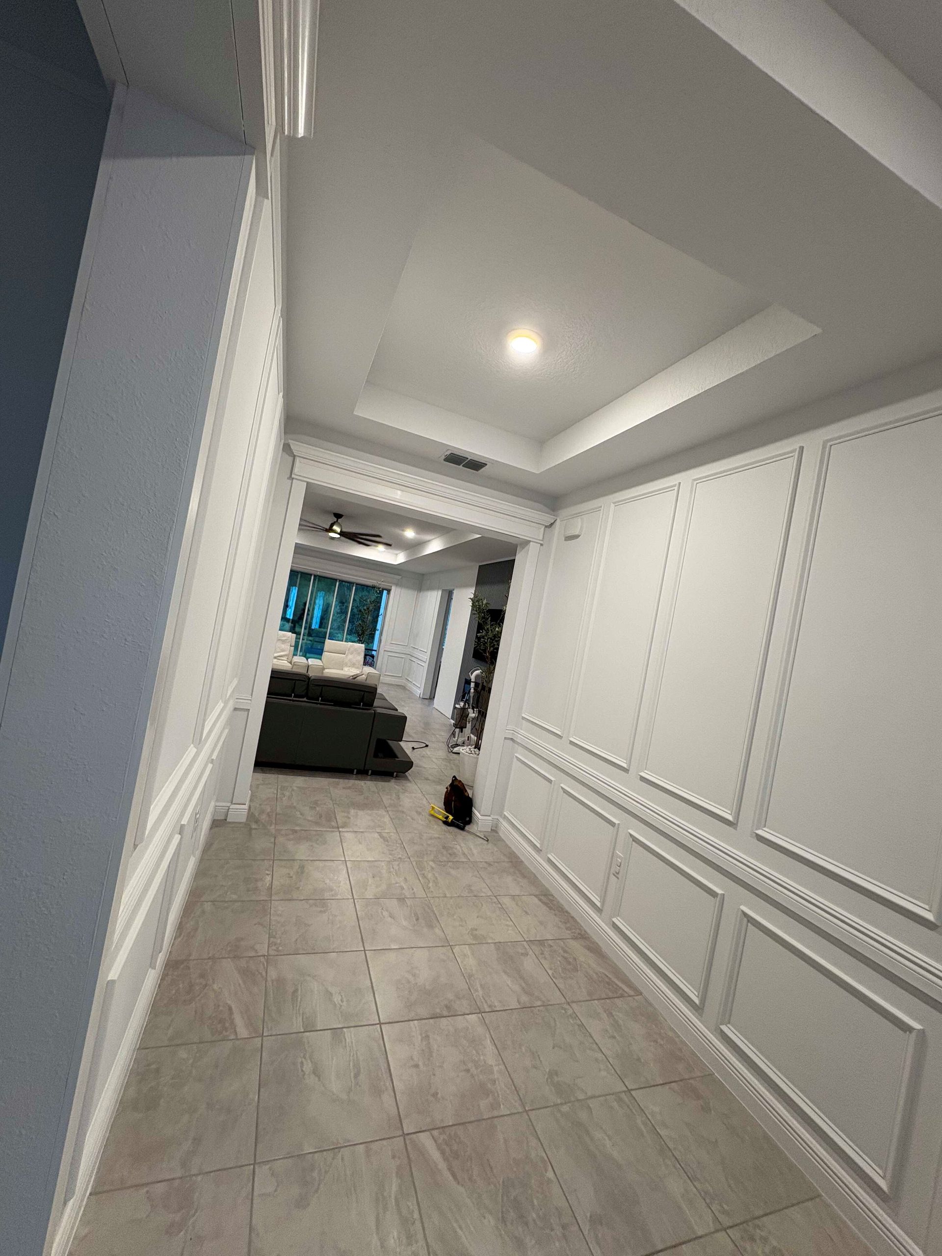 Bright hallway with white paneled walls, tiled floor, and a glimpse of a living room at the far end.
