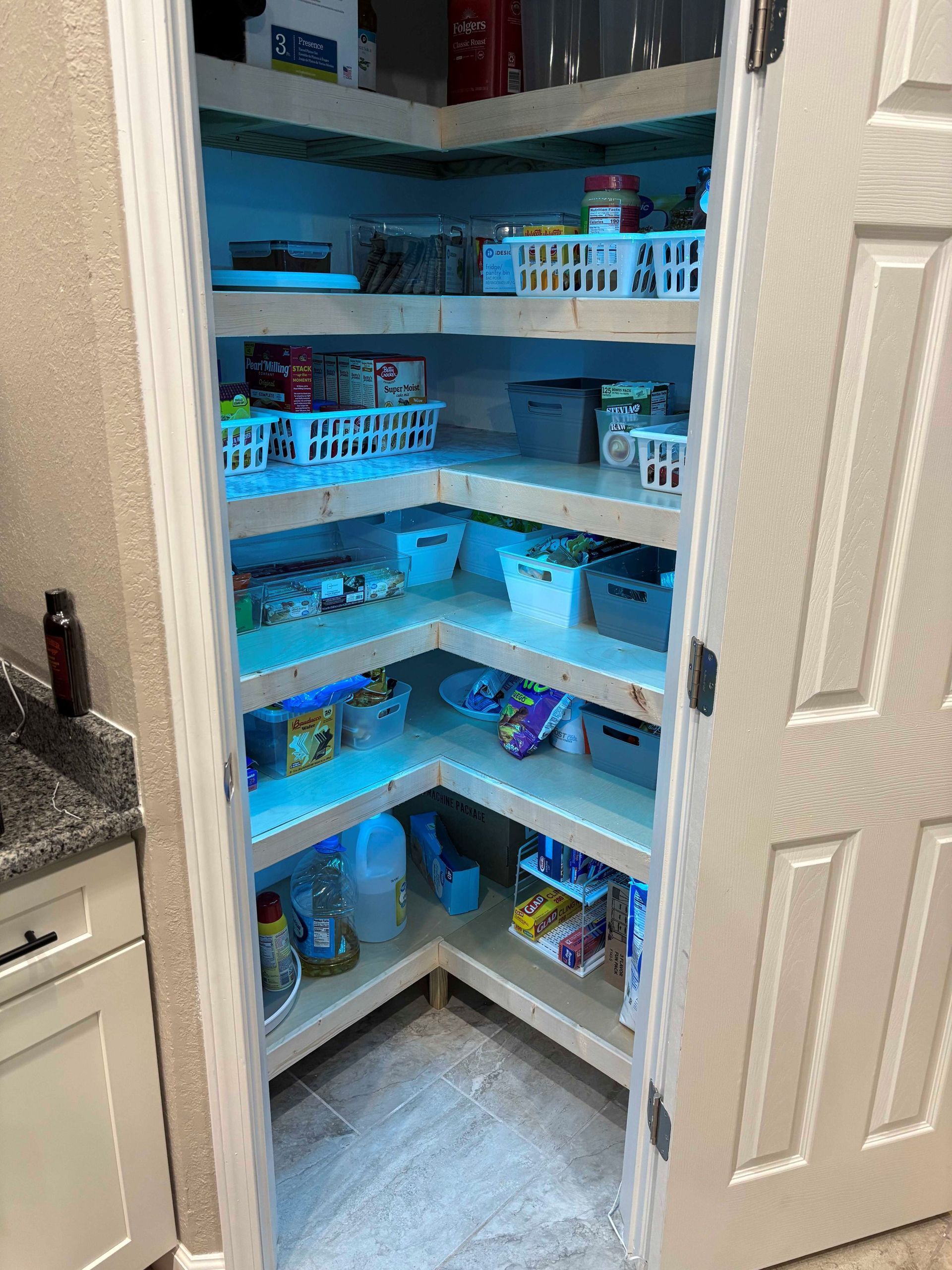 Corner pantry with blue-lit shelves holding baskets and snacks, viewed through a partially open door
