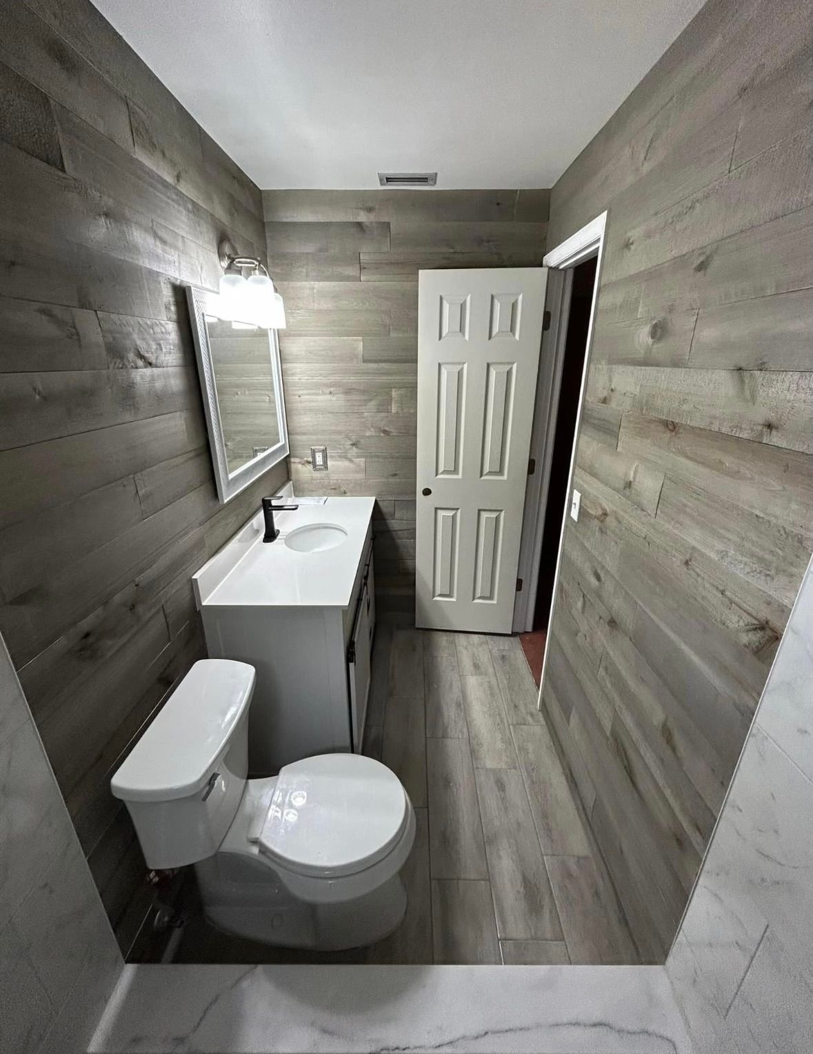 Narrow bathroom with gray wood walls, white toilet and sink, and an open door at the end.